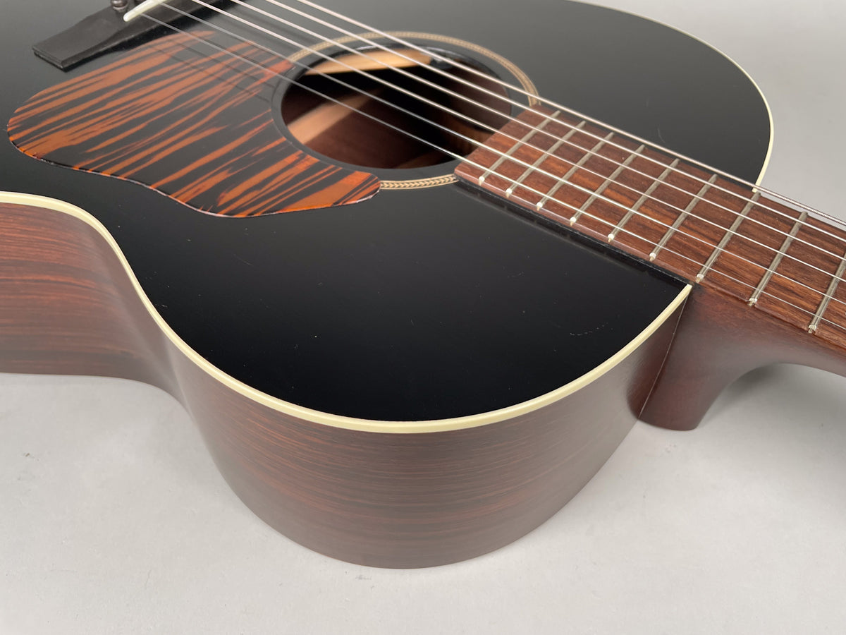 Close-up of the black acoustic guitar's body, showing the dark wood sides, a striped pickguard, and the fretboard extending from the neck.