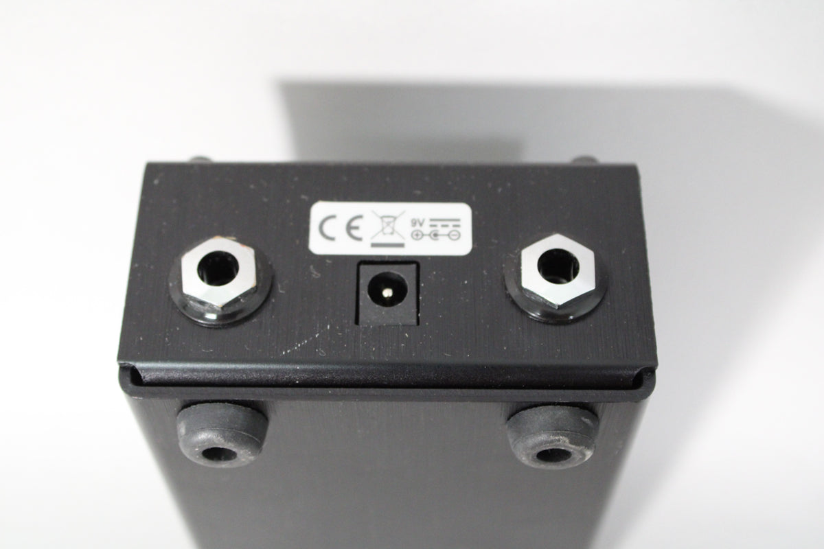 Top-down view of the black pedal's rear, showing two silver-hexagonal jack inputs, a central power input, and a white label.