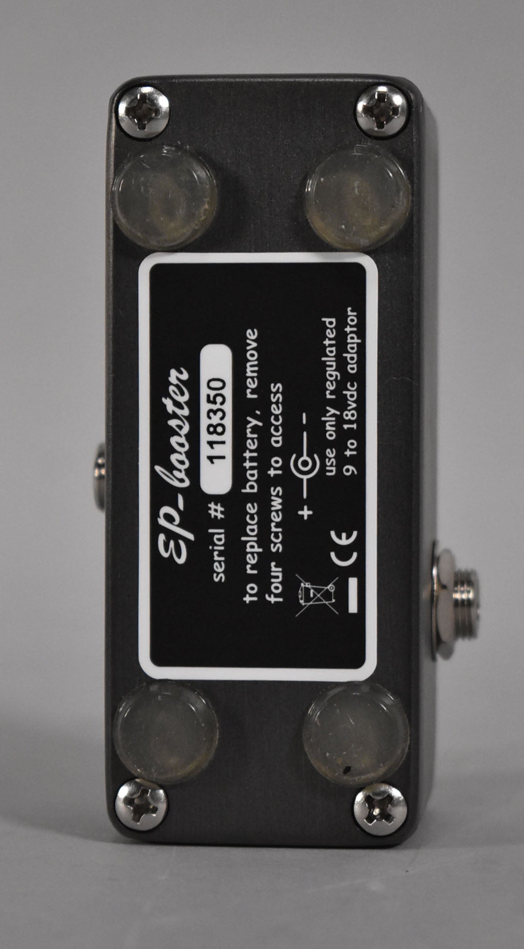 The dark gray mini pedal is viewed from the bottom, showing four clear rubber feet, four silver screws, and a black label.