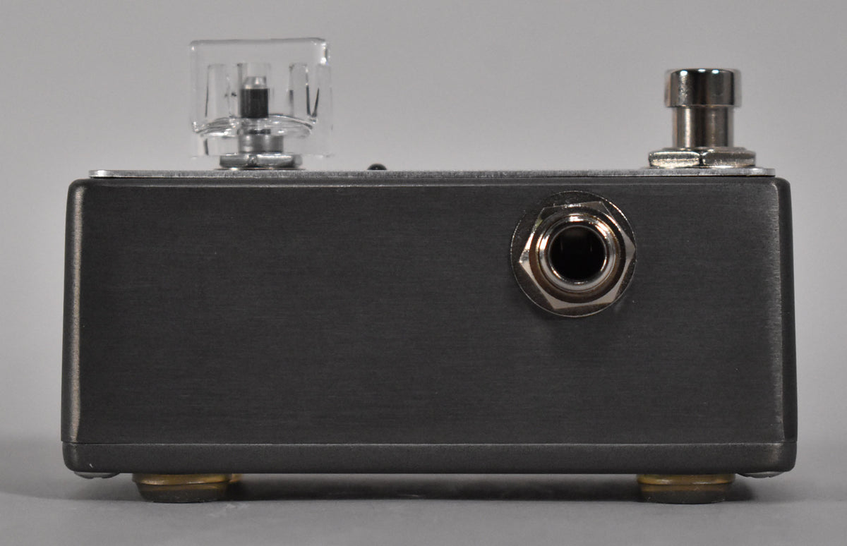 The dark gray pedal is viewed from the side, showing a clear knob, a chrome input jack, and a silver footswitch.