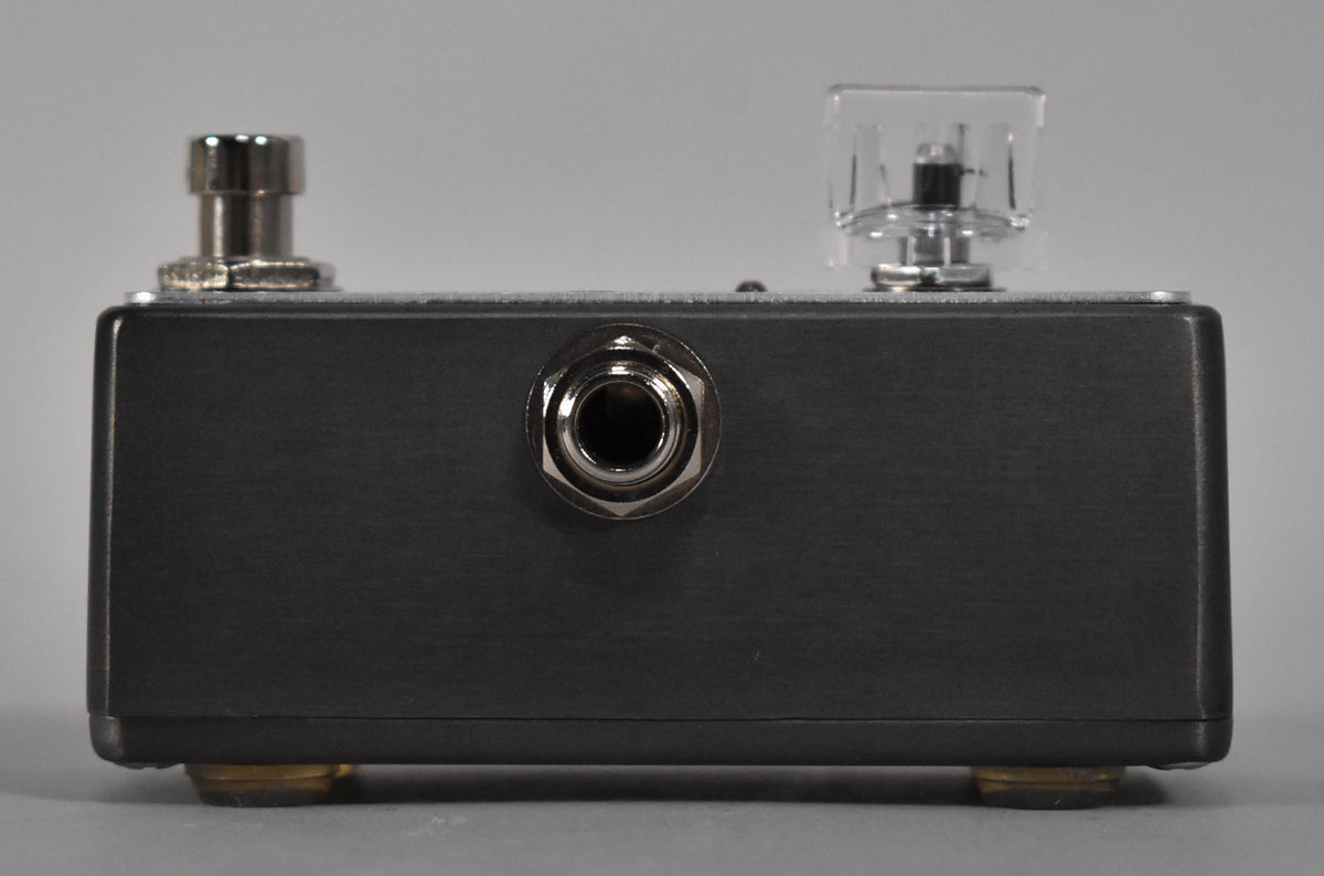 The dark gray pedal is viewed from the side, showing a silver input jack, a footswitch, and a clear control knob.