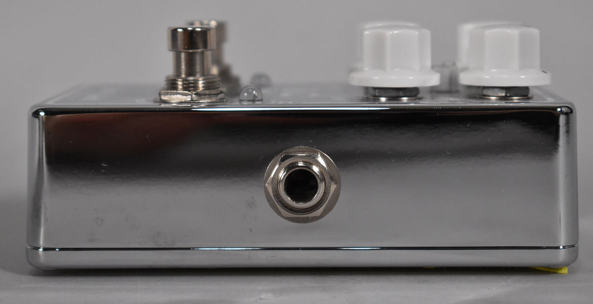 The chrome-finished pedal is viewed from the side, showing a single input jack and the bottom edge of two white control knobs.