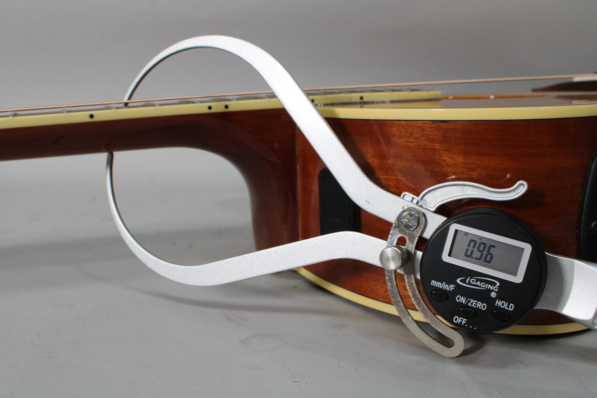 A digital caliper measures the neck relief of the acoustic guitar, showing 0.96mm, against its dark brown body and light binding.