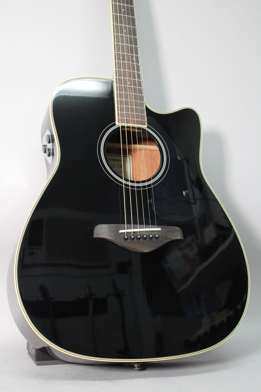 The glossy black acoustic-electric guitar is angled, showing its cutaway, soundhole, dark bridge, and side-mounted preamp controls.