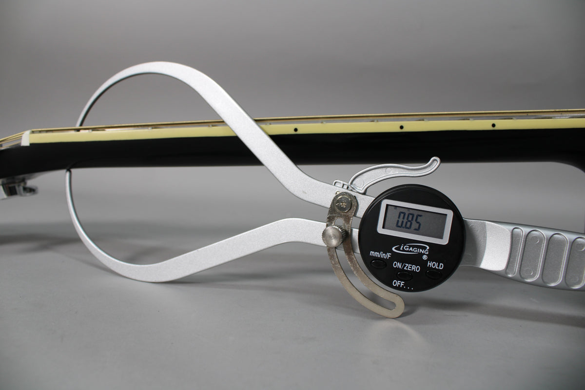 A digital caliper measures the black guitar's neck relief at 0.85mm, with the fretboard and strings visible from the side.