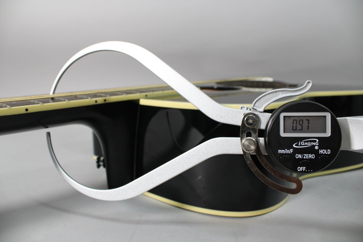 A digital caliper measures the black guitar's action at the 12th fret, showing 0.97mm, with a cream binding visible.