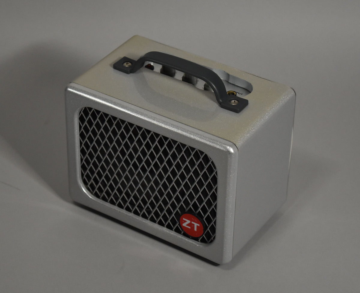 A small, silver-grey textured amp with a black diamond-patterned grille, a red circular logo, and a black top handle.