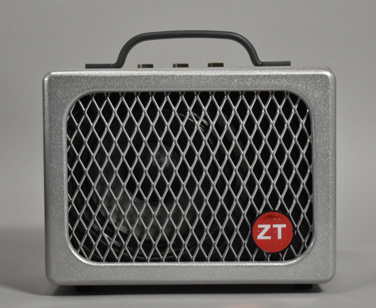 Front view of a silver sparkle amp with a diamond-patterned metal grille, black handle, and a red circular logo in the lower right.