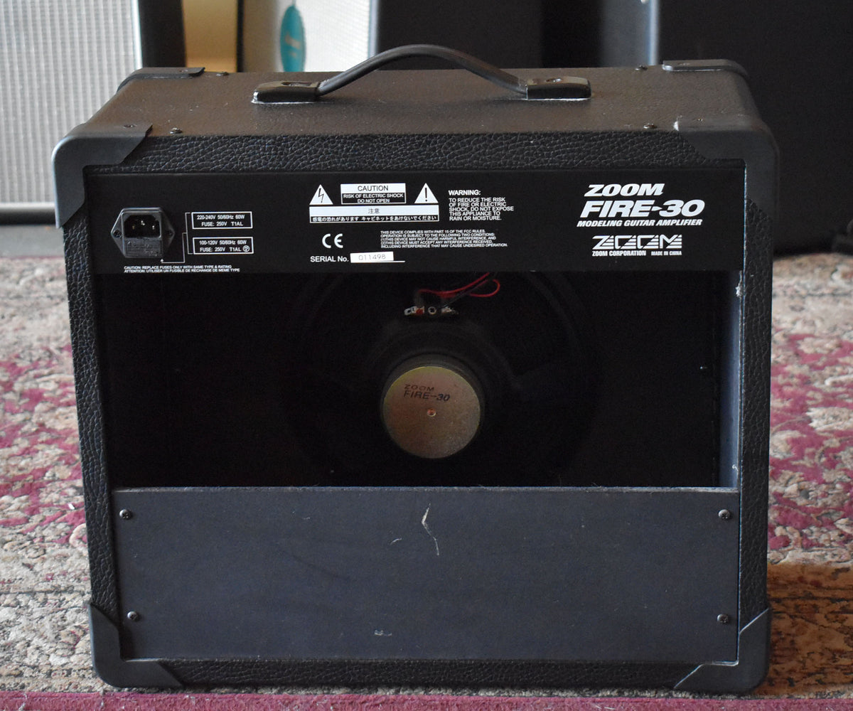 Rear view of a black textured amp, showing the power input, warning labels, and a visible speaker with a gold center.