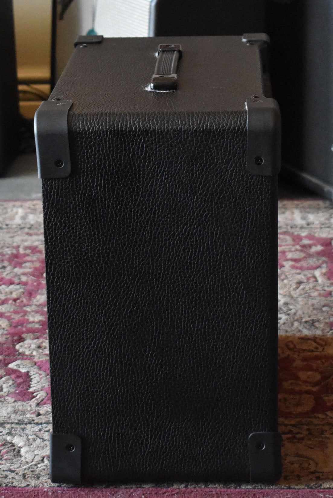 The black textured amp is seen from the side, with a top handle and four black corner protectors visible.