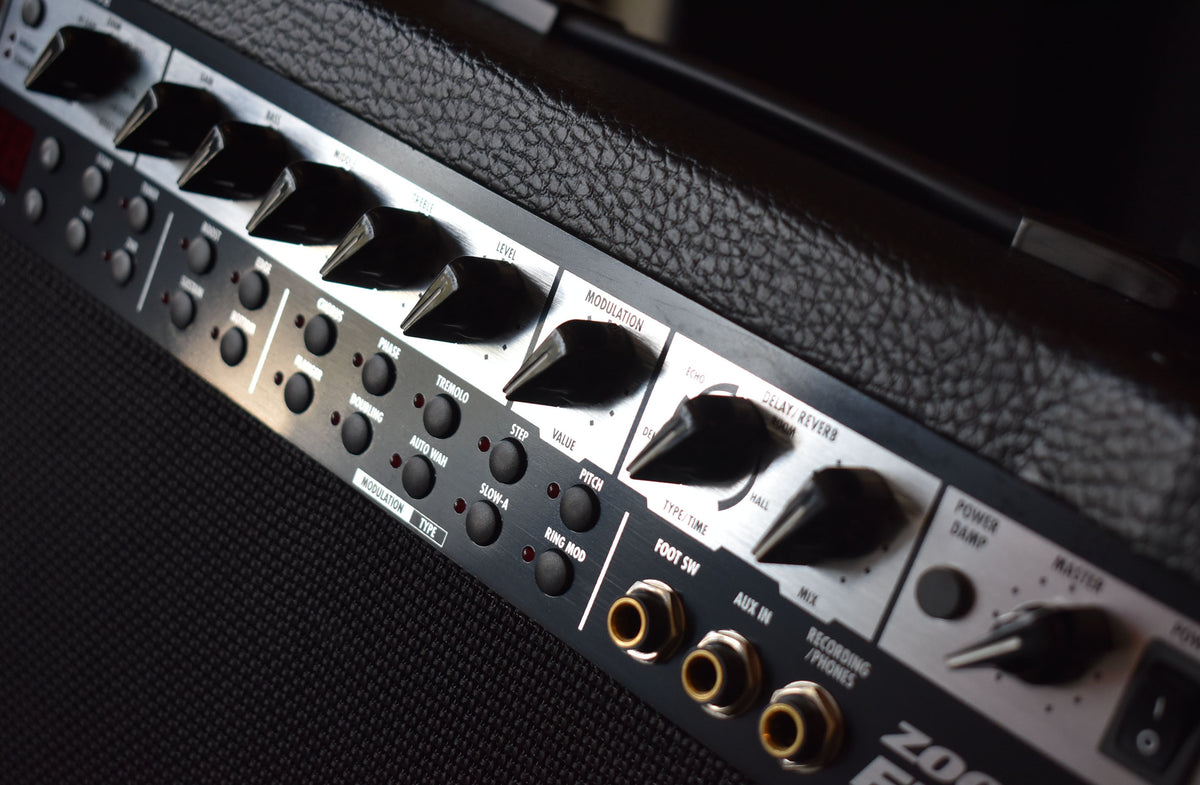 Close-up of the amp's black control panel, angled from the left, showing numerous black knobs and buttons, and three gold-colored input jacks.