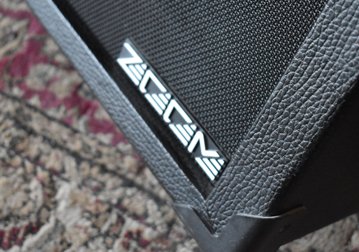 Close-up of the amp's upper right corner, showing the black textured tolex, speaker grille, and a silver logo badge.
