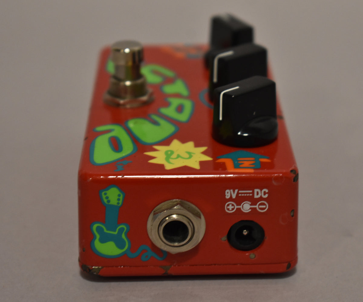 A red guitar pedal with hand-painted green letters, a guitar, and a sun, showing input/output jacks and 9V DC power.