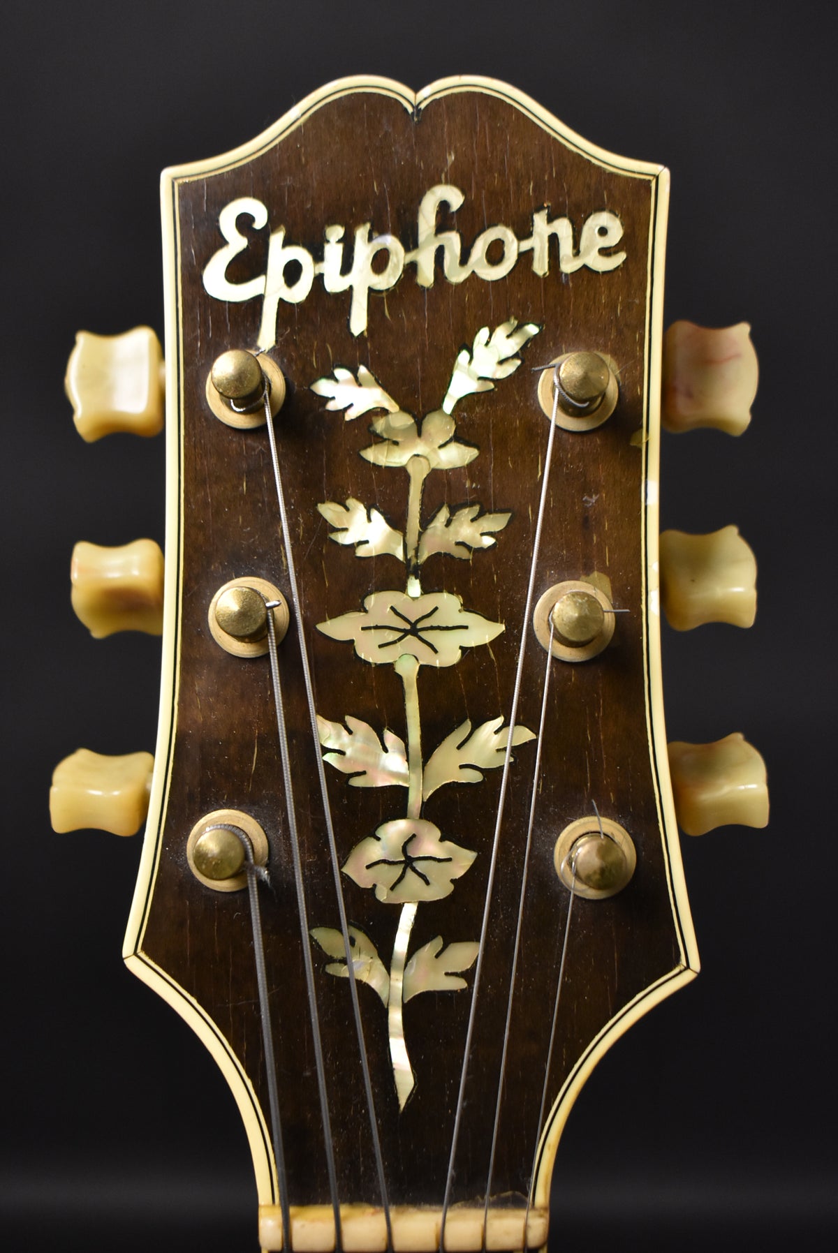 Close-up of the dark brown headstock, featuring ornate mother-of-pearl inlay and six cream-colored tuning pegs with brass posts.