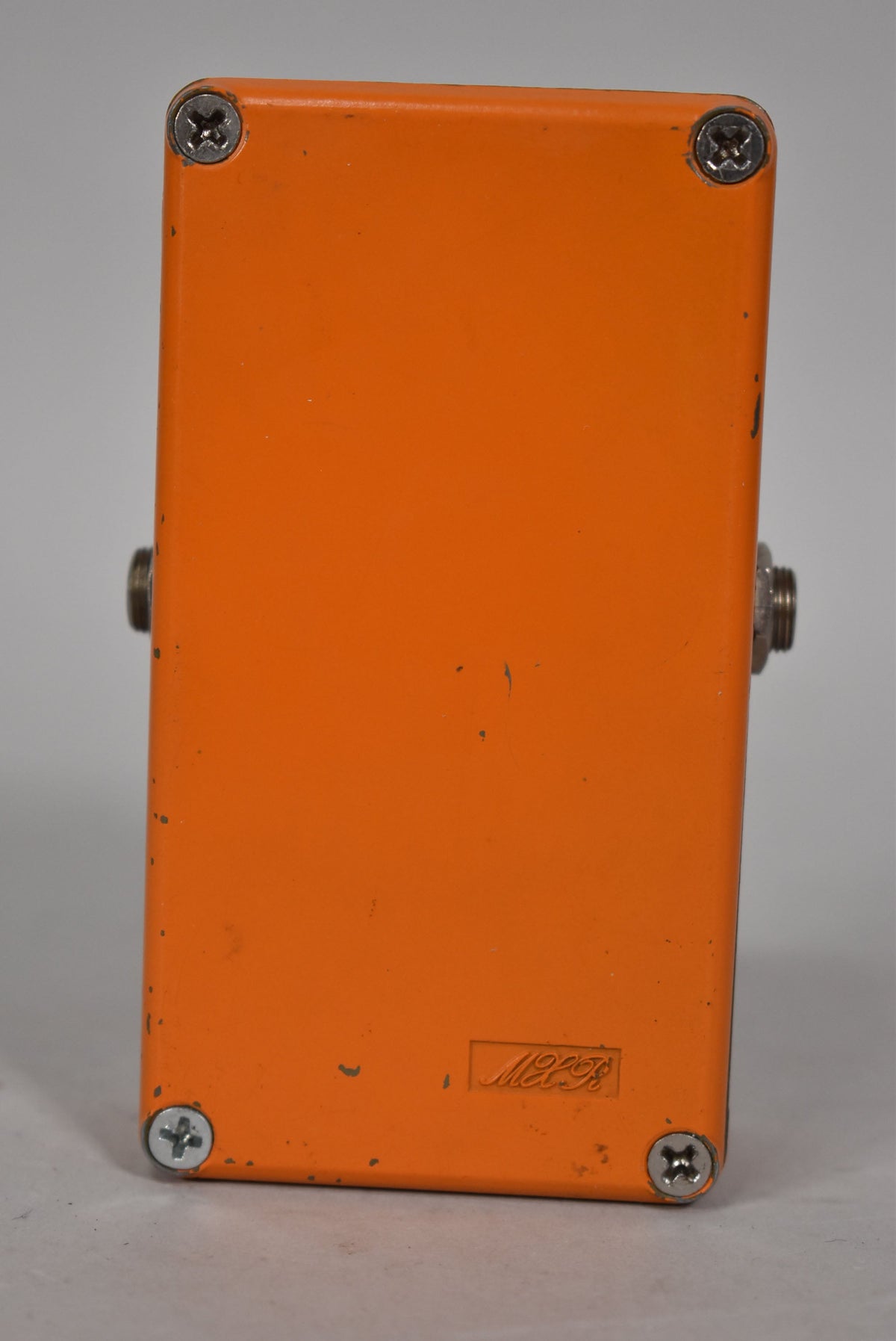 The orange pedal's back is shown, with four silver screws, two side jacks, and a faint rectangular logo near the bottom.