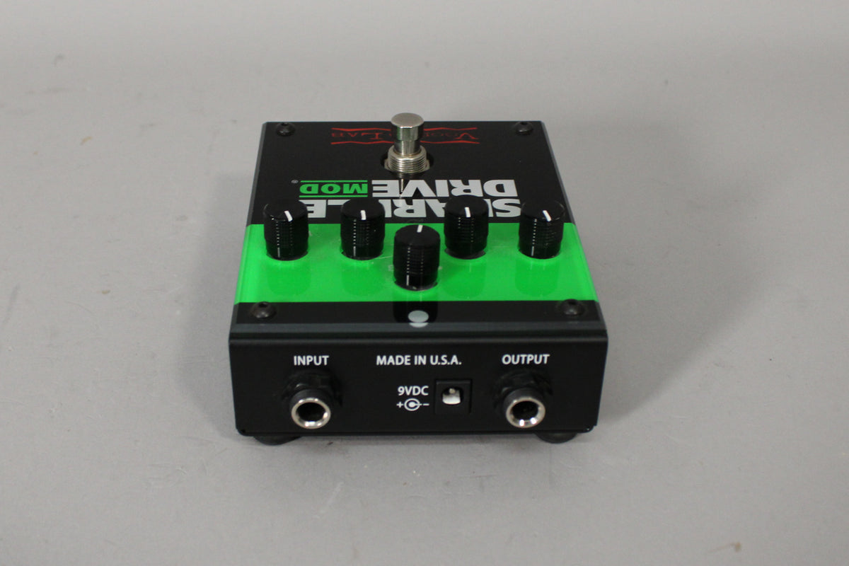 A black and green pedal, viewed from the front, with five black control knobs, a silver footswitch, and input/output jacks.