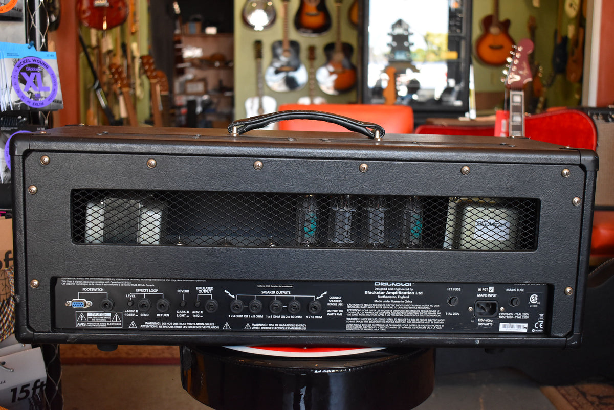 Rear view of the black textured amp head, showing a metal grille revealing five glowing tubes and a full panel of inputs and outputs.