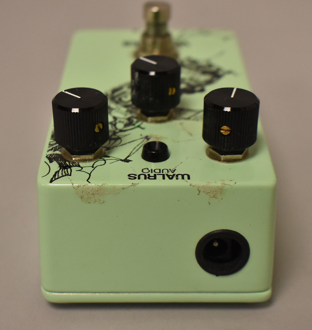 A green effect pedal, viewed from the front, shows three black knobs, a power input, and wear on the top and front edges.