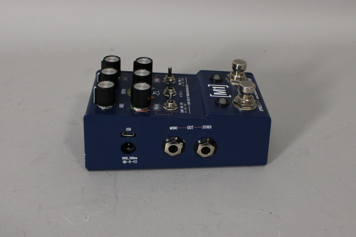 Dark blue pedal, angled from the left, showing six black knobs, three silver toggle switches, two footswitches, and stereo output jacks.