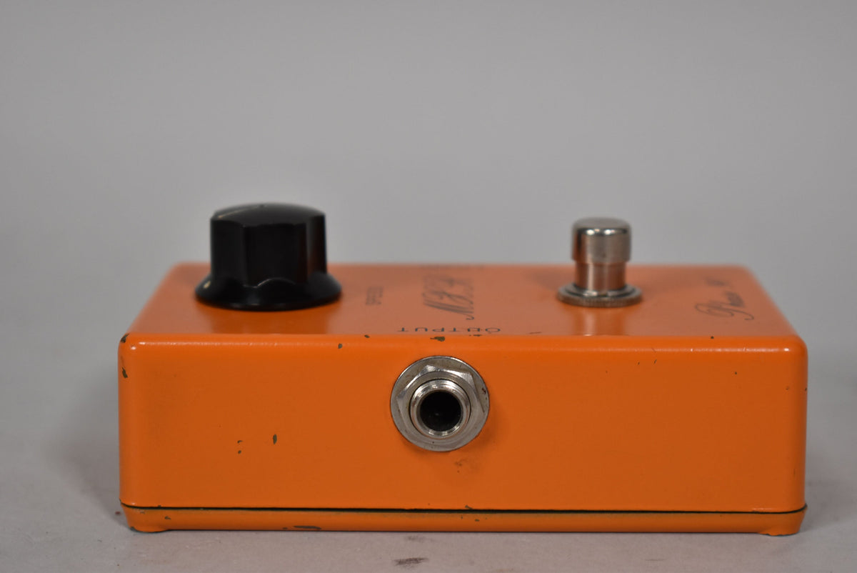The orange pedal is viewed from the front, showing the output jack, a black knob, and a silver footswitch, with visible paint chips.
