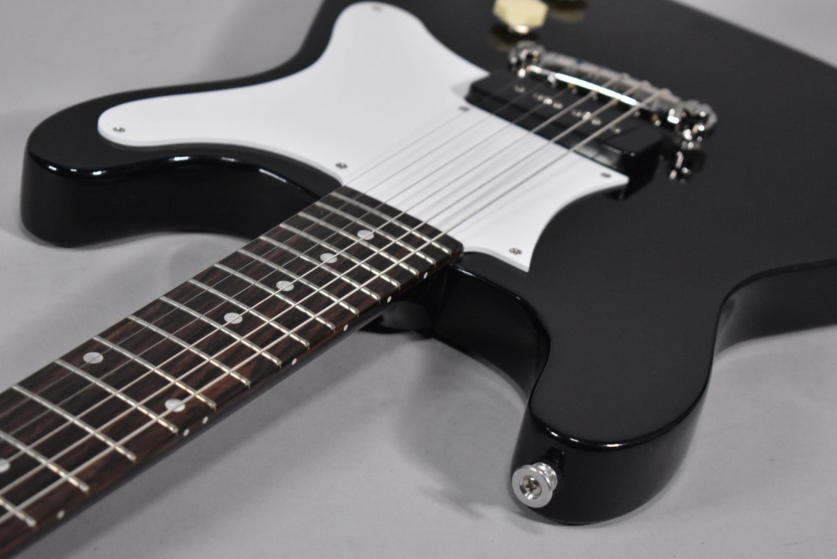Close-up of a glossy black electric guitar with a white pickguard, chrome bridge, and dark fretboard with dot inlays.