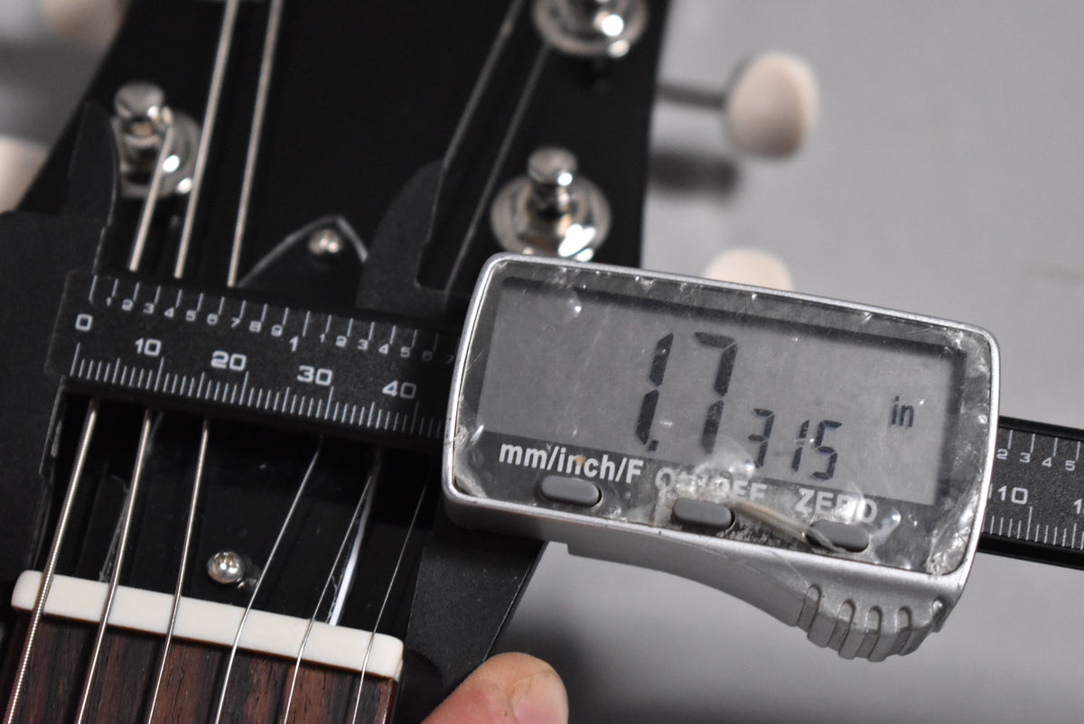 A digital caliper measures the guitar's nut width at 1.7315 inches, with the headstock, tuners, and strings visible.