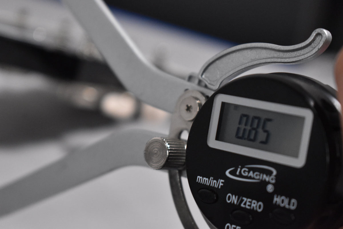 Close-up of a digital caliper measuring 0.85, with its silver arms and black display visible against a blurred background.