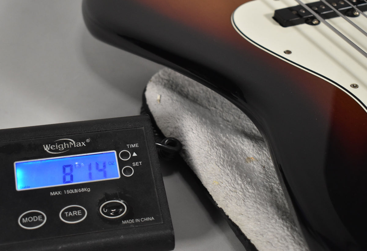 The bass guitar's lower body, in 3-tone sunburst, rests on a grey case lining, with a scale displaying "8 LB 14 OZ" in the foreground.