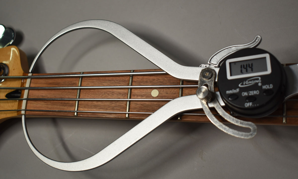 A digital caliper measures the bass's fretboard, showing a reading of 1.44, with a light-colored headstock visible.