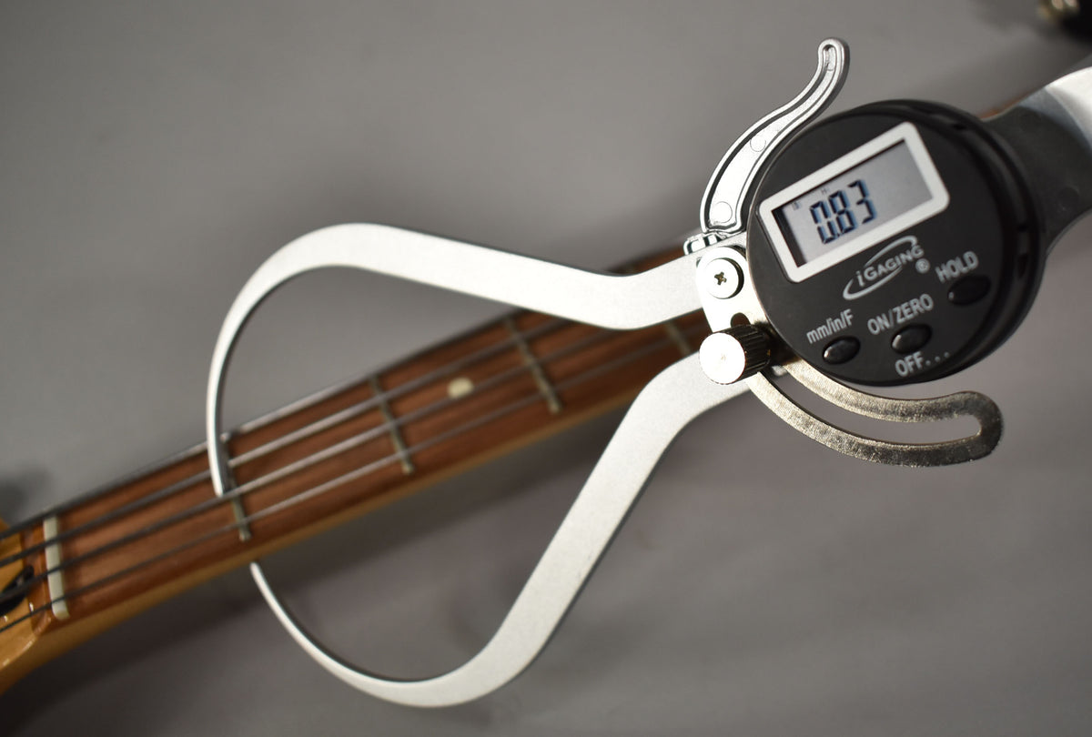 A digital caliper measures the bass's fretboard and strings, displaying "0.83" on its screen.