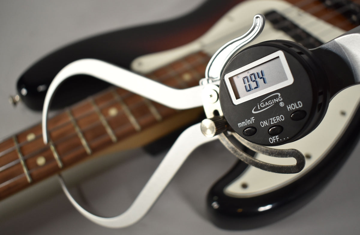A digital fret height gauge shows "0.94" over the bass's rosewood fretboard, with its sunburst body blurred in the background.