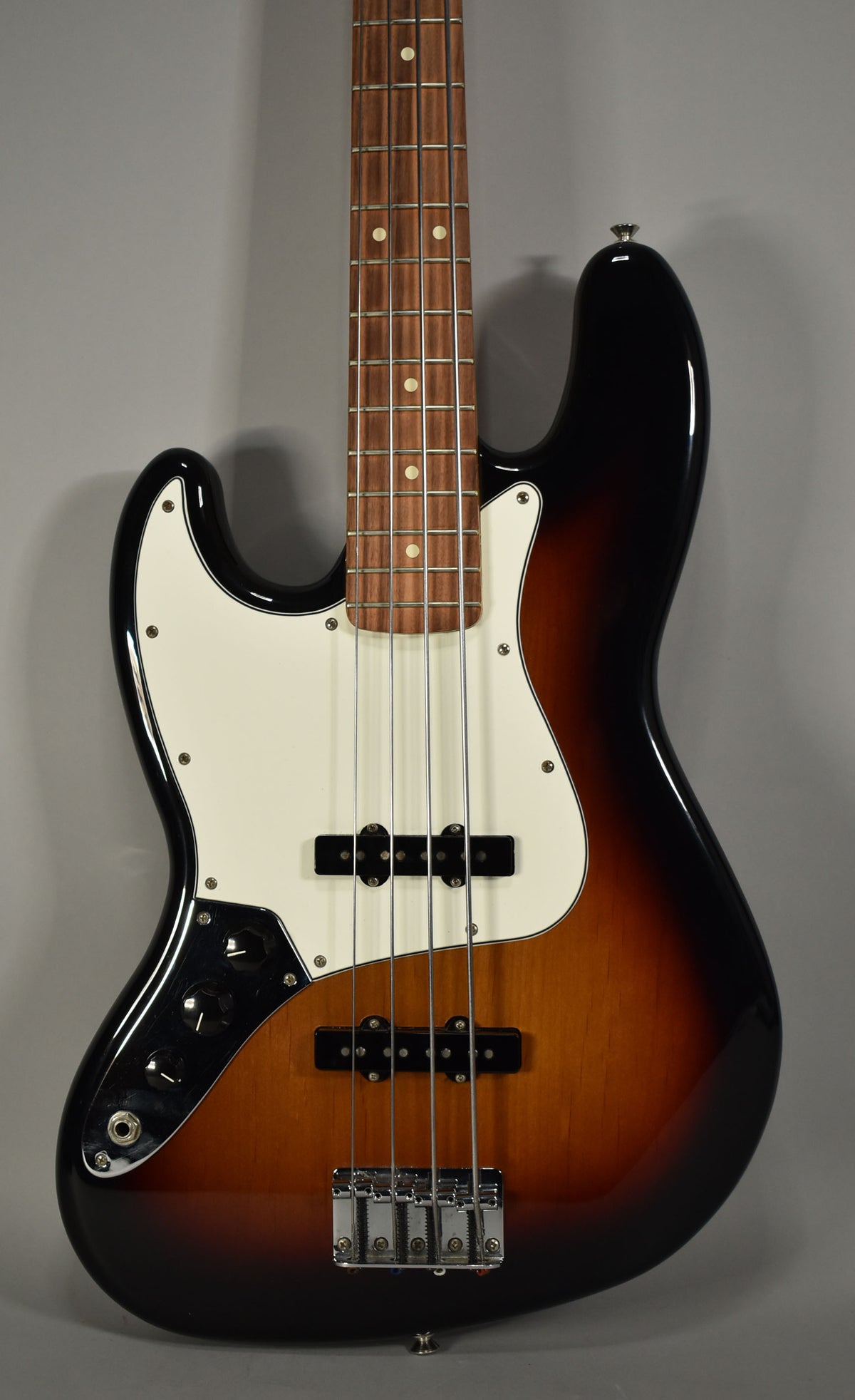 The left-handed Jazz Bass, in 3-Tone Sunburst, is angled slightly right, showing its white pickguard, two pickups, and chrome bridge.