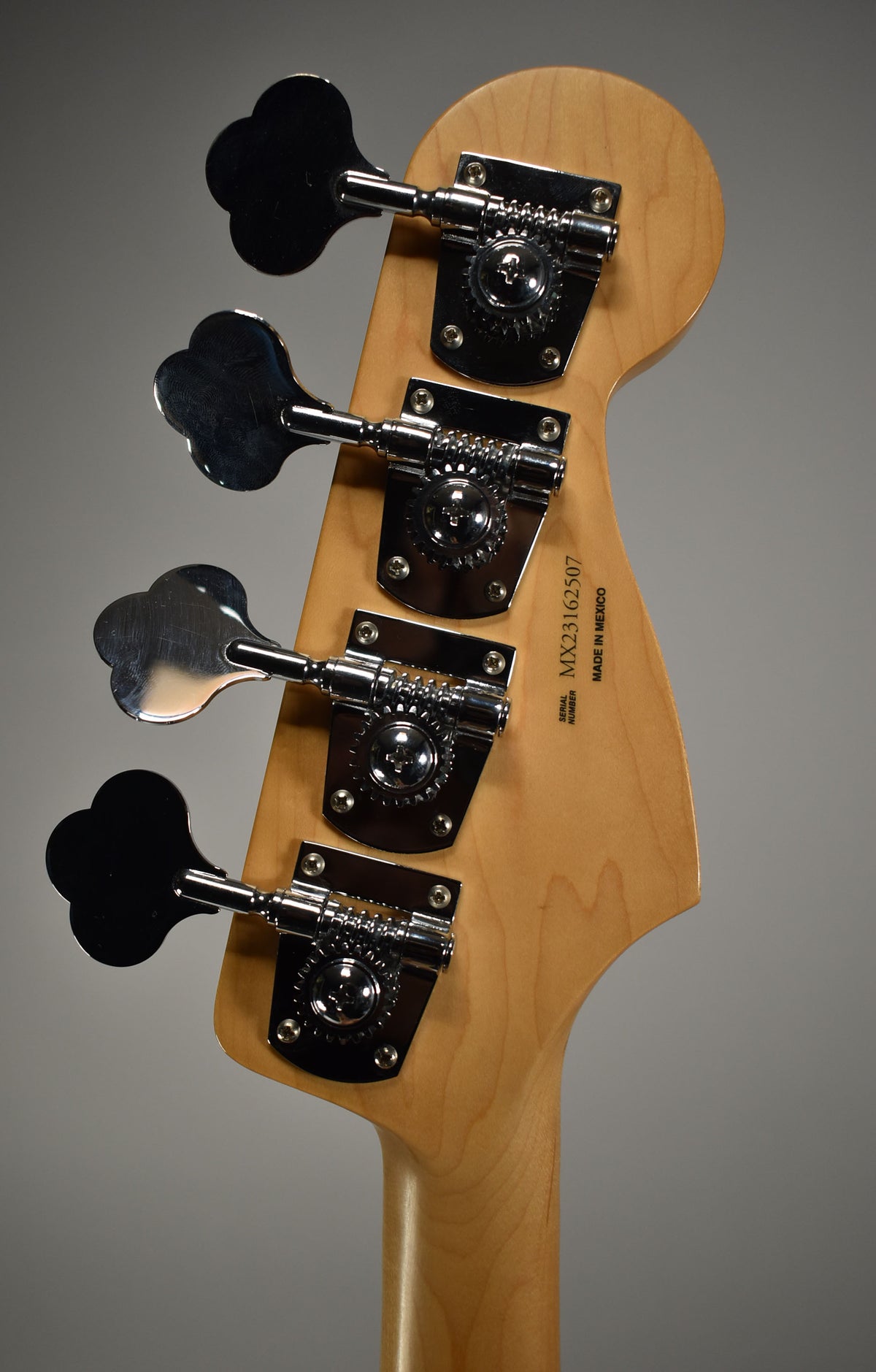Rear view of the bass headstock and top of the neck, showing four clover-leaf tuners with chrome hardware on light wood.