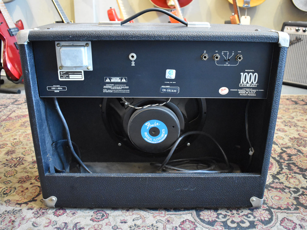 Rear view of a black textured combo amp, showing the open back cabinet with a single speaker, various input jacks, and a power transformer.