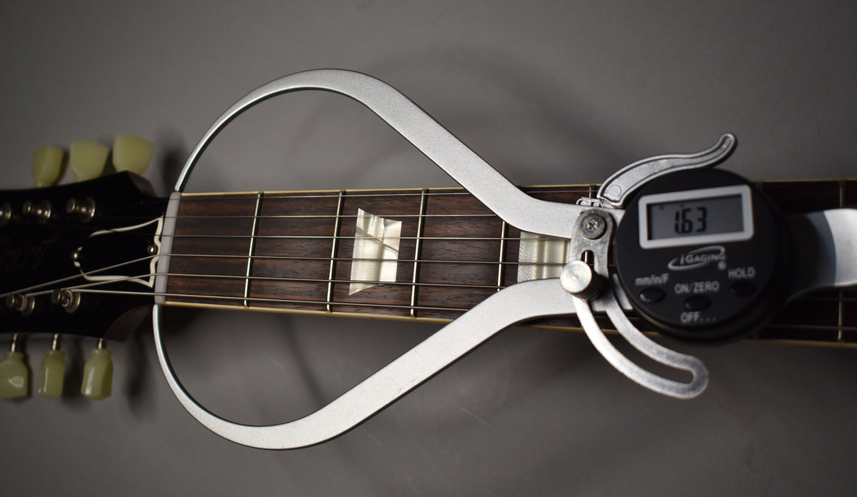 A digital string action gauge measures 1.63mm on the dark rosewood fretboard with trapezoid inlays, near the bone nut and headstock.