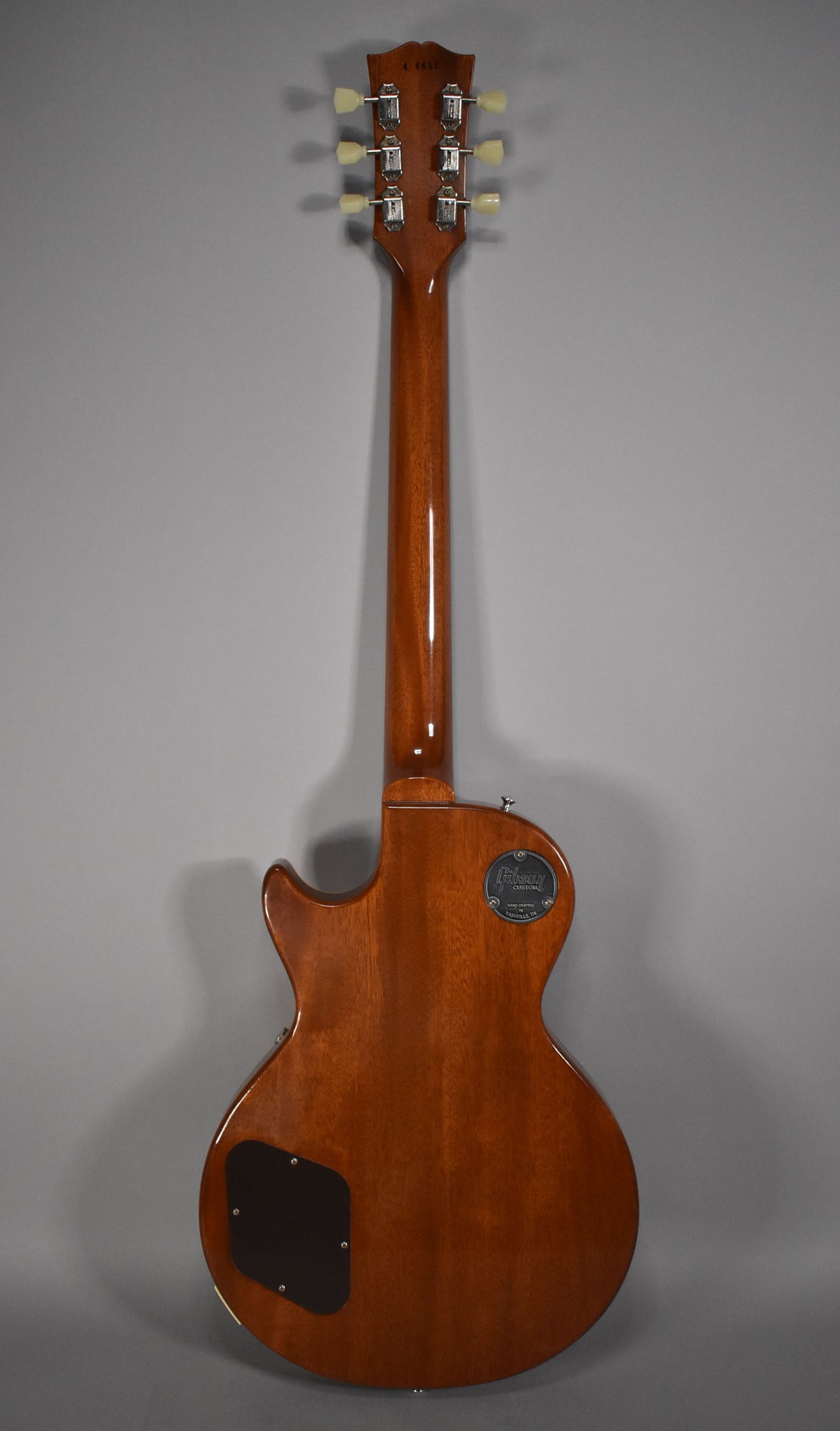 Full rear view of a glossy, medium-brown guitar with a dark control cavity cover and a round, metallic backplate near the neck joint.