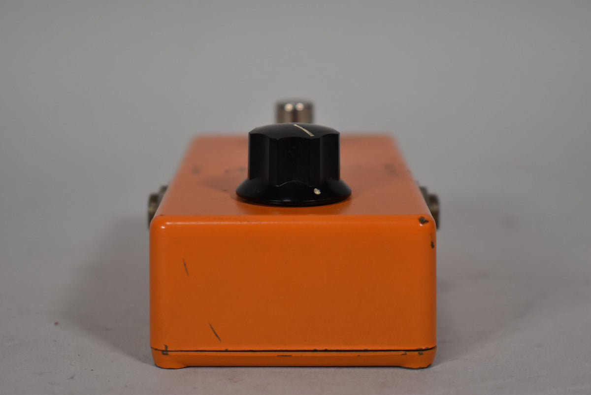 The orange pedal is viewed from the front, showing the black control knob with a white indicator line and some paint chips.