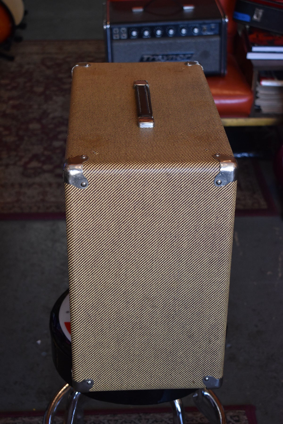 The tweed-covered cabinet is angled from above, showing the top and right side, with a dark brown leather handle and chrome corner protectors.