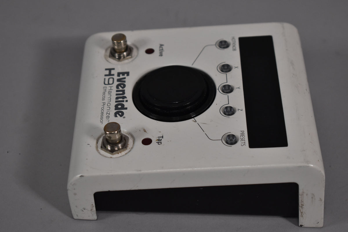 Top-down view of a white pedal with a large black central knob, two footswitches, and a small black screen.
