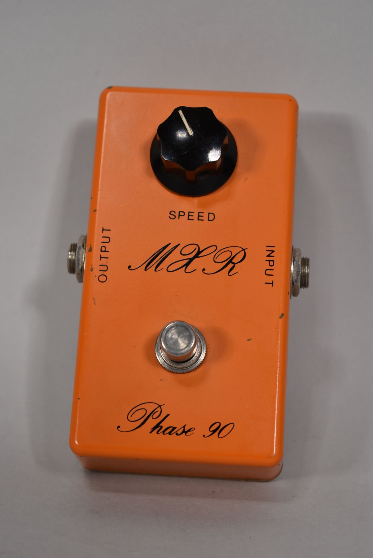 An orange rectangular effects pedal, viewed from above, with a single black knob, a silver footswitch, and two silver jacks.