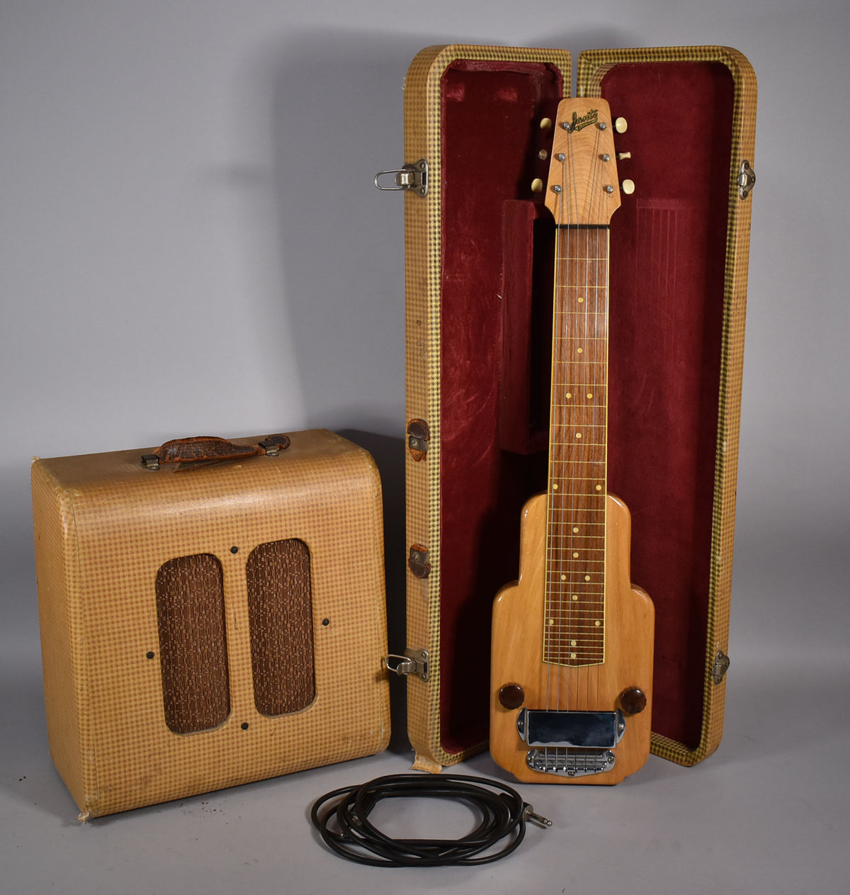 A light wood lap steel with a chrome bridge and two dark brown knobs, resting in its red-lined tweed case, next to a matching tweed amplifier.