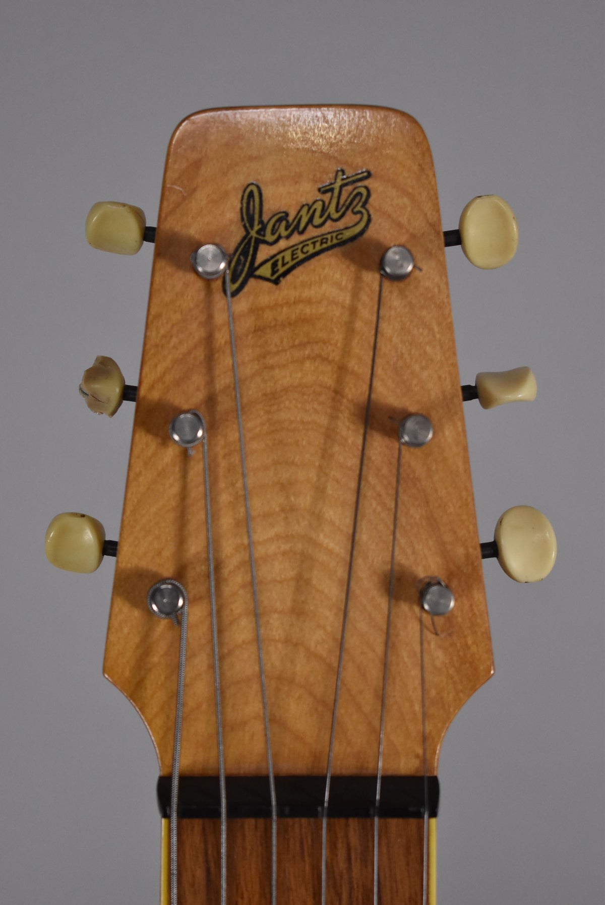 Close-up of the light brown wooden headstock with six strings, six cream tuning pegs (one chipped), and a black and yellow logo.