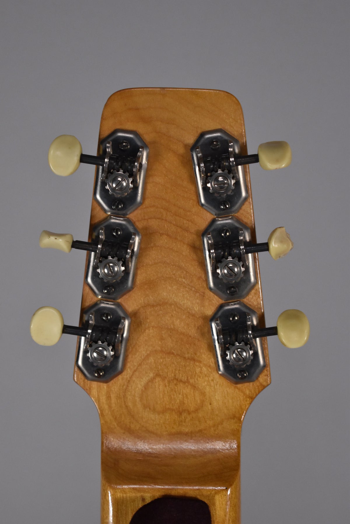 The back of a light-brown wooden headstock with six chrome tuners and cream buttons, one of which is chipped.