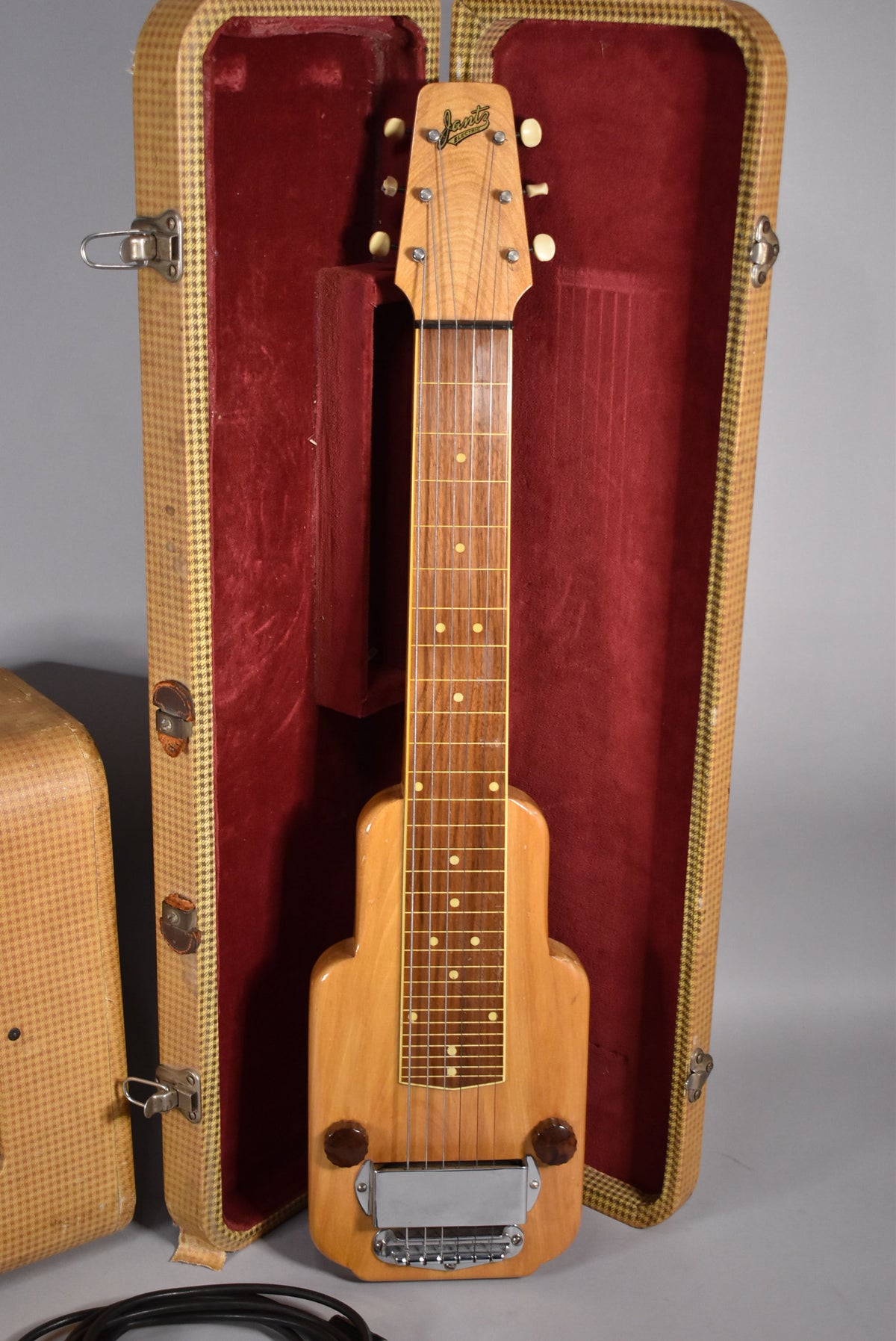 A natural wood lap steel guitar with a chrome bridge and two dark brown control knobs, resting in a red velvet-lined tweed case.