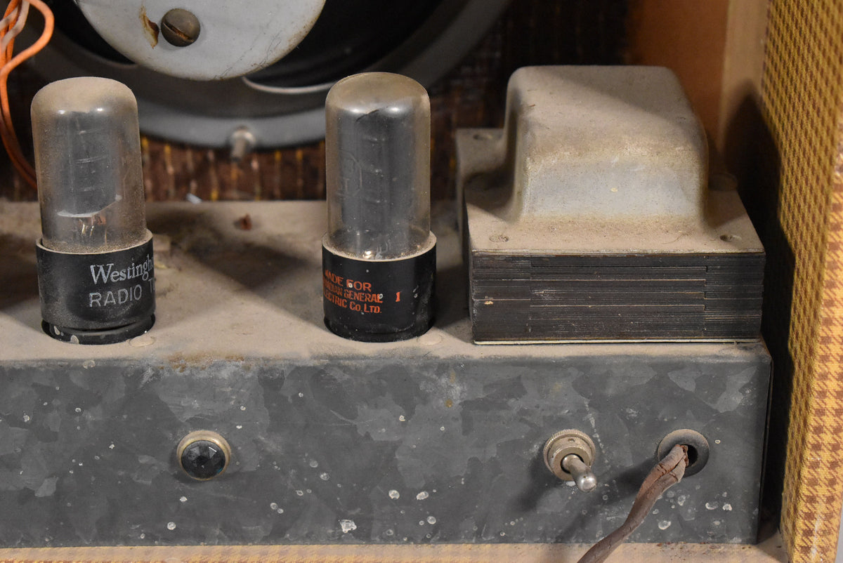 Close-up of the amplifier's dusty, speckled gray metal chassis, showing two vacuum tubes, a transformer, a black knob, a toggle switch, and a frayed power cord.
