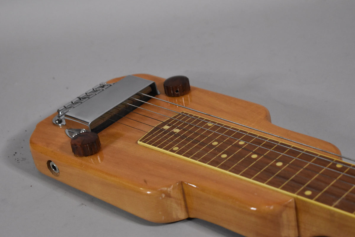 Close-up of a light-brown lap steel's body, showing two dark brown knobs, a chrome bridge cover, and a fretboard with dot inlays.