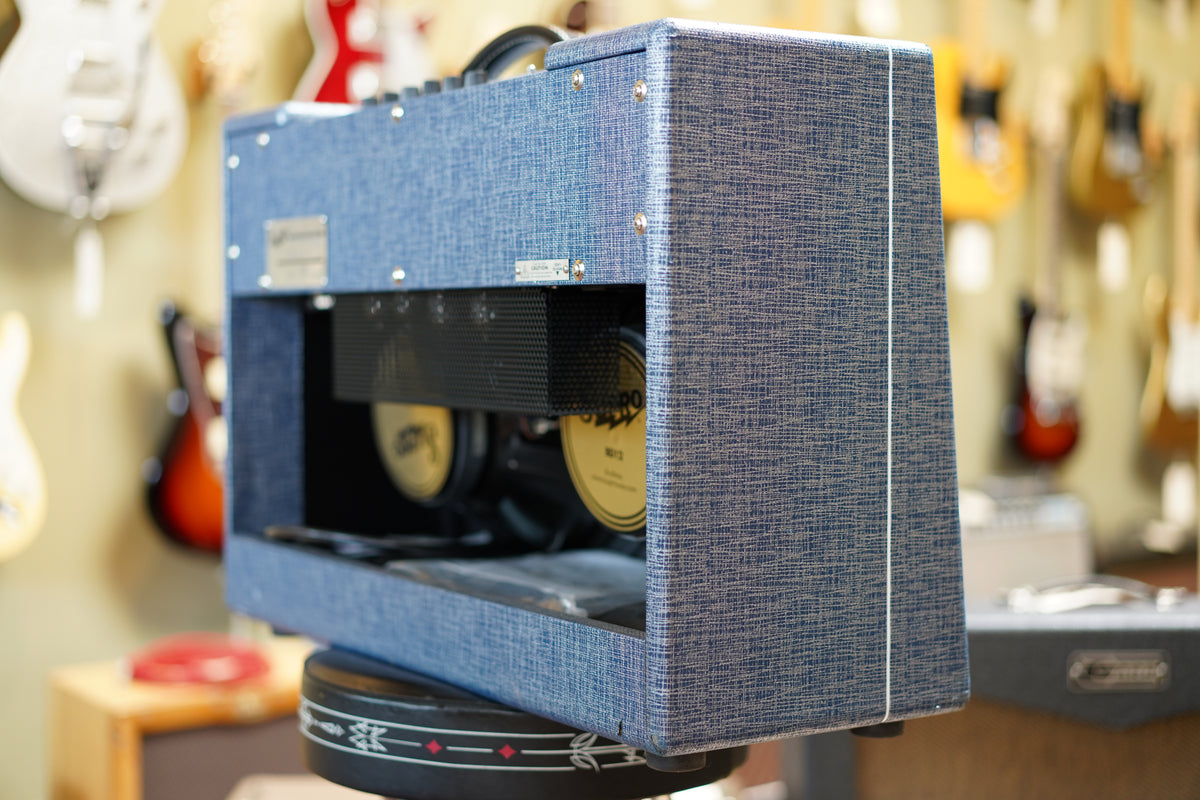 The blue tweed amp is angled to show its right side and rear, revealing two gold-coned speakers behind a black mesh panel.