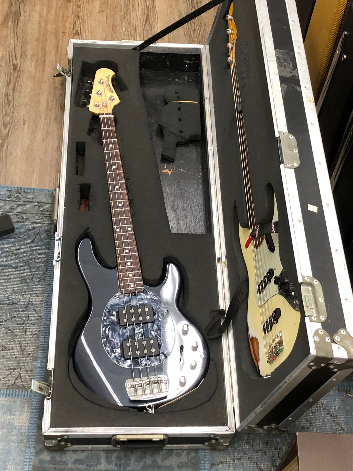 An open black flight case with silver trim, containing a dark blue bass with a pearloid pickguard and a cream-colored bass.