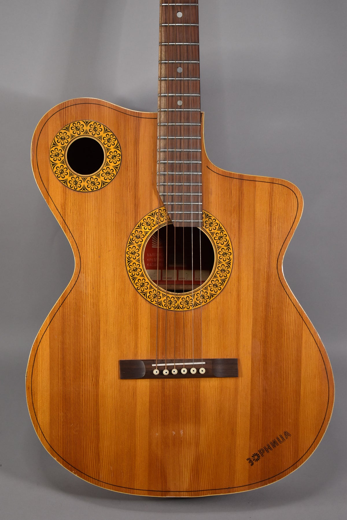 The natural finish acoustic guitar's body and neck are framed, showing two soundholes with ornate yellow and black floral rosettes.