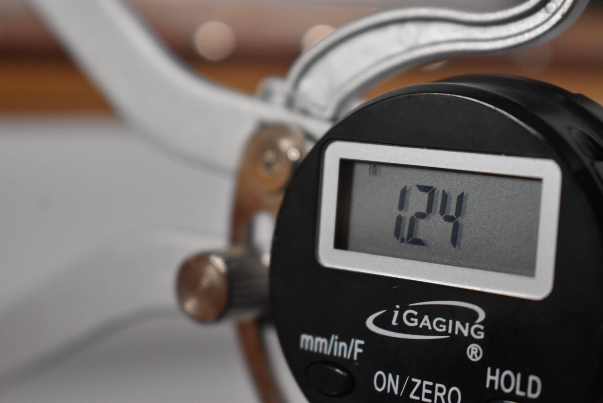 Close-up of a digital caliper measuring string height at 1.24mm, with the guitar's natural finish neck blurred in the background.